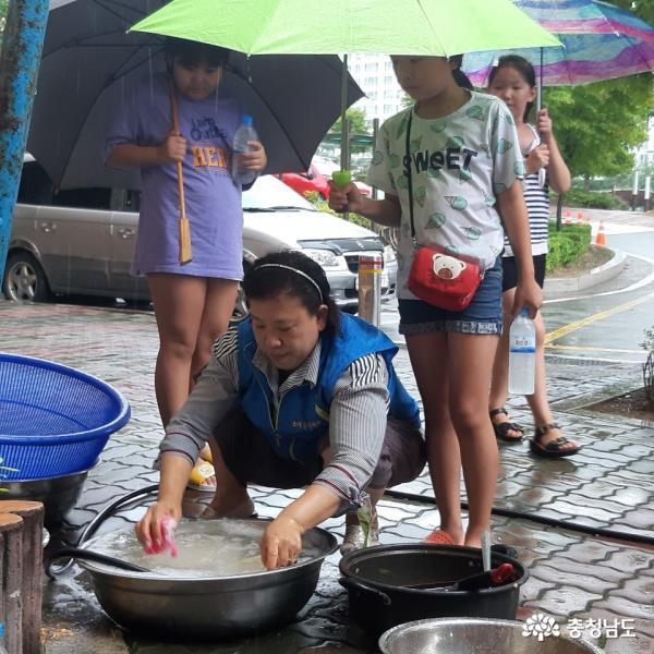  소은희 부녀회장이 비를 맞으며 뒷마무리 설거지 하는 모습을 보던 어린 학생들이 우산을 받쳐주고 있다.   