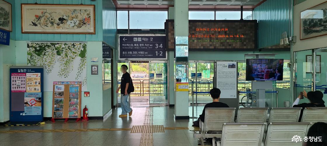 강경역 내부 사진. 출입구 너머로 연두색 풍경이 들어오고, 역사 내에는 대기석에 앉아 기차를 기다리는 사람들이 있습니다.