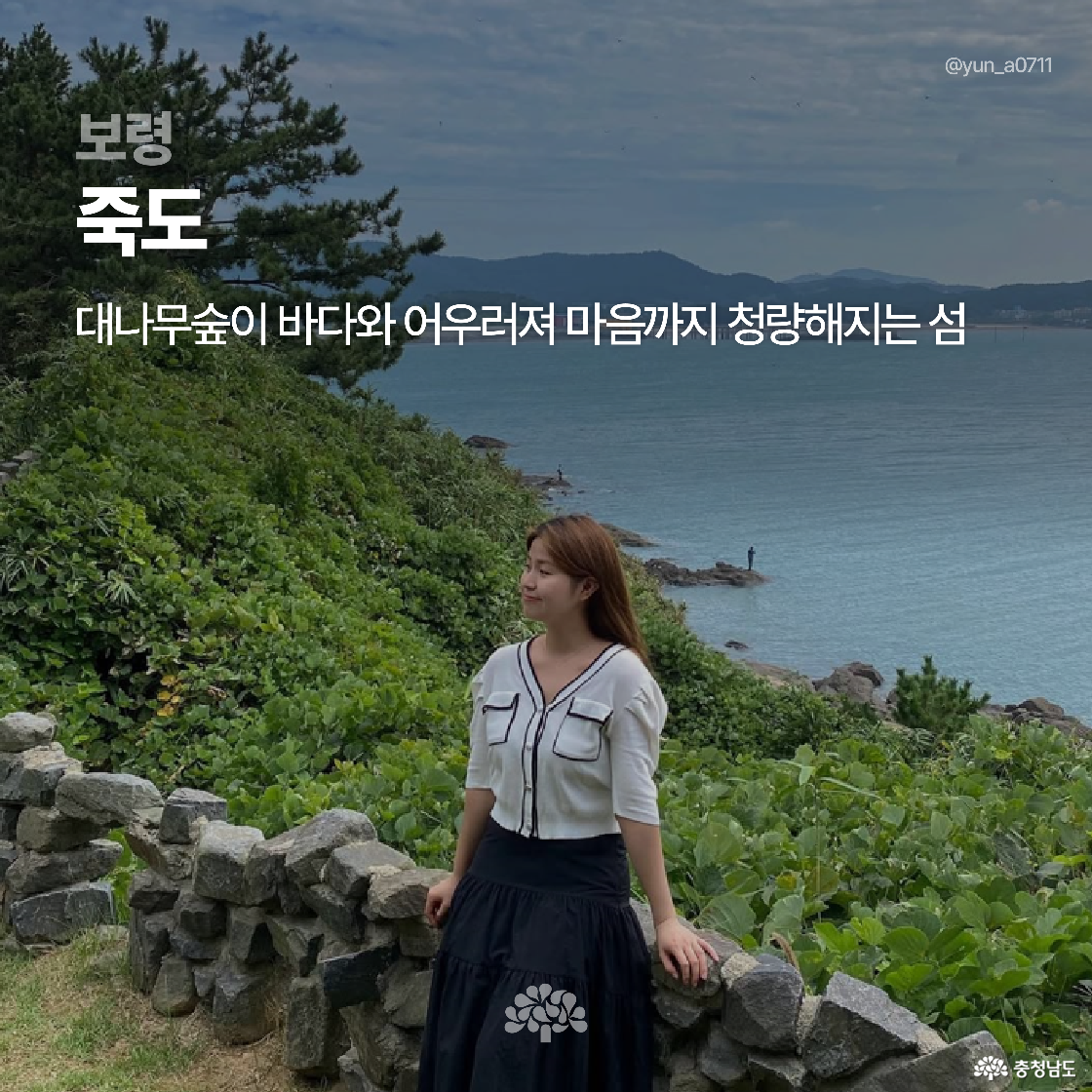 보령 죽도의 해안가가 배경으로, 대나무숲과 바다가 조화를 이루며 초록빛 풍경이 펼쳐져 있습니다. 돌담 앞에는 흰 블라우스와 검정 스커트를 입은 여성이 미소를 지으며 서 있는 모습이 담겨 있습니다.
