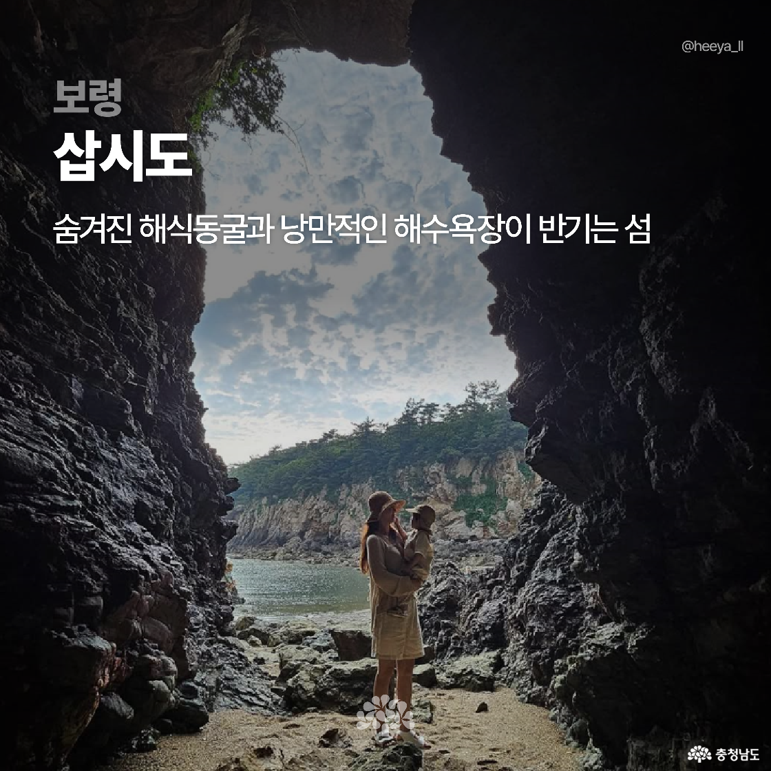 해식동굴 안에서 바깥쪽 바다를 바라보는 장면으로, 모자와 원피스를 입은 여성이 아이를 안고 있습니다. 자연적으로 생긴 암벽 틈새를 통해 파란 하늘과 숲이 보이며 ‘보령 삽시도’의 낭만적인 분위기가 느껴집니다.