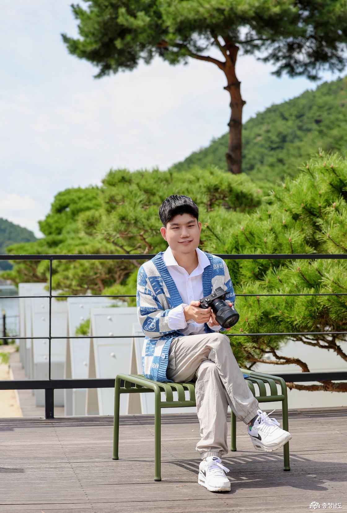푸른 소나무와 산을 배경으로 전성민 씨가 카메라를 들고 야외 의자에 앉아 환하게 미소 짓고 있습니다. 