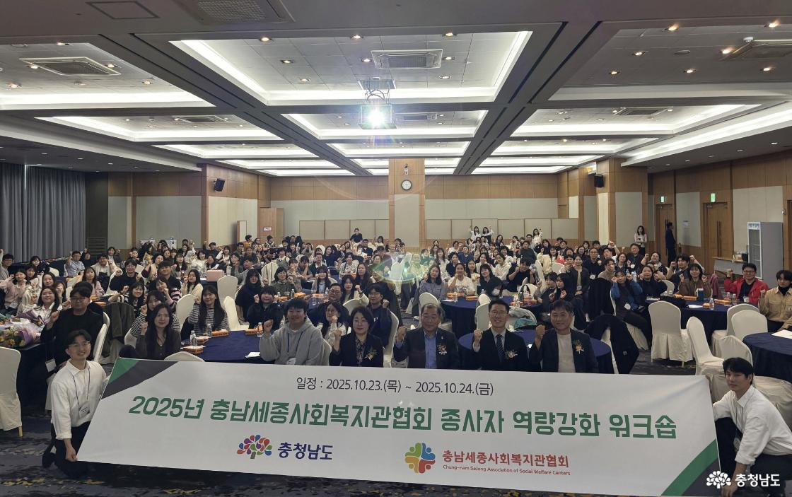2025년 충남세종사회복지관협회 종사자 역량강화 워크숍 행사 단체 사진. 실내 회의실에서 많은 참석자들이 테이블에 앉아 카메라를 향해 웃으며 손을 들어 인사하고 있다. 앞줄에는 협회 관계자들이 행사 현수막을 들고 포즈를 취하고 있으며, 현수막에는 ‘2025년 충남세종사회복지관협회 종사자 역량강화 워크숍, 일정: 2025.10.23(목)~10.24(금), 충청남도, 충남세종사회복지관협회’라고 적혀 있다.