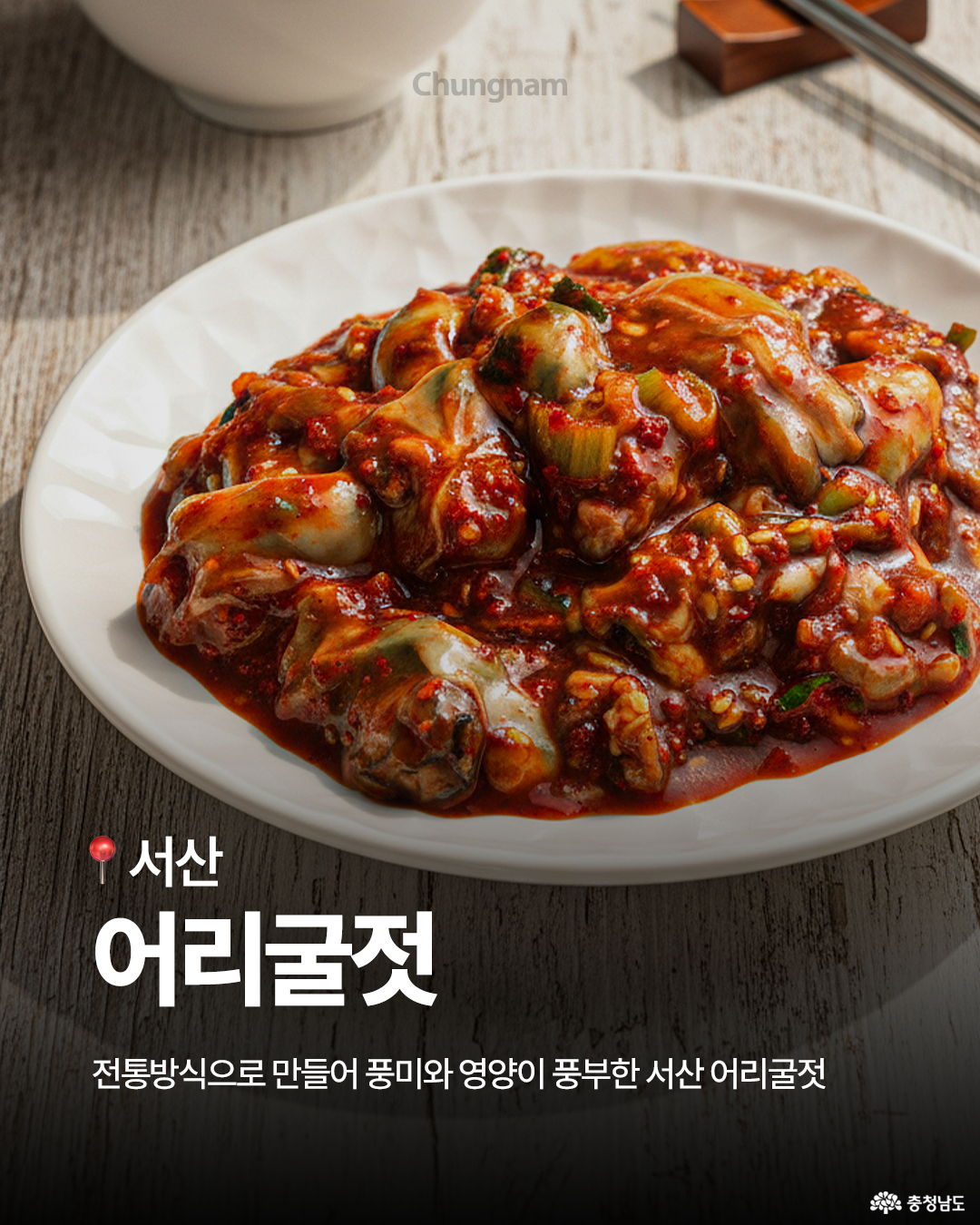 흰 접시 위에 빨갛게 양념된 어리굴젓이 수북하게 담겨 있으며, 굴살과 고춧가루 양념의 질감이 생생합니다. 뒤로는 나무 젓가락과 밥그릇이 함께 놓여 있어 한식 반찬의 분위기를 살립니다. 하단에는 “서산 어리굴젓 – 전통방식으로 만들어 풍미와 영양이 풍부한 서산 어리굴젓”이라는 설명이 있습니다.