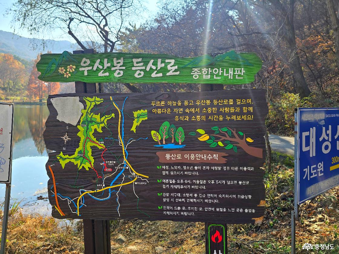 우산봉 등산로 종합안내판&rsquo;이라는 제목의 나무질 안내판이 서 있다. 안내판에는 등산로 지도가 색깔로 표시되어 있으며, 초록색 산길과 파란색&middot;노란색 길이 서로 연결된 모습이 그려져 있다. 오른쪽에는 등산로 이용 안내 수칙과 그림 아이콘이 함께 적혀 있다. 안내판 뒤로는 가을빛 산책로와 저수지가 흐릿하게 보인다