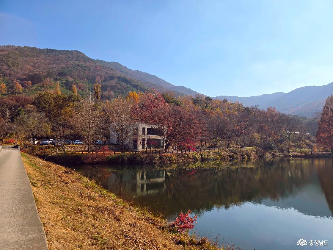 고요한 저수지 수면에 가을빛 산자락과 나무들이 선명하게 반사되어 있다. 물가 뒤편에는 흰색 2층 건물과 여러 대의 차량이 나무 사이로 보이고, 주변 나무들은 붉은색&middot;노란색&middot;갈색 등 가을빛으로 물들어 있다. 멀리 산 능선이 겹겹이 펼쳐져 부드러운 분위기를 이루고, 왼쪽에는 산책길이 이어져 있다