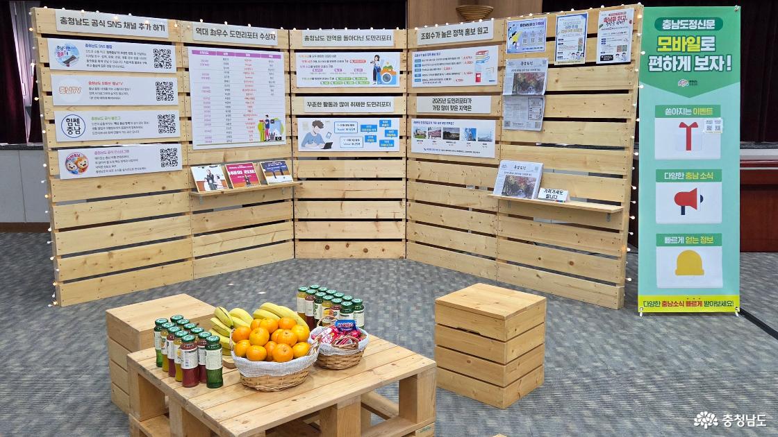 2025 충청남도 도민리포터 성과공유회 전시 부스2(충남 공식 SNS 채널 안내와 역대 최우수도민리포터 수상자 명단, 올해 활약한 도민리포터 명단 등이 게시된 나무 전시월, 그 앞으로 참석자들을 위한 다과가 정성스럽게 차려져 있습니다.