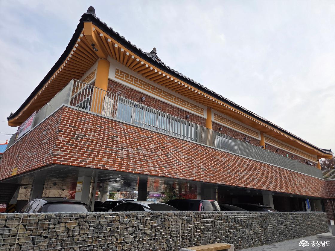 붉은 벽돌과 전통 기와지붕이 조화를 이룬 홍주천년문화체험관 건물의 웅장한 측면 모습입니다. 건물 하부에는 넉넉한 주차 공간이 마련되어 있고, 돌을 채워 만든 독특한 담장이 인상적입니다.