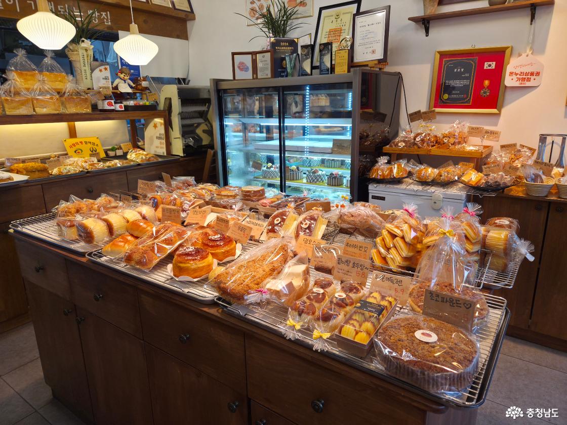 가운데 매대에 다양한 빵이 가득 진열되어 있고, 벽쪽으로 케이크 냉장고와 귀여운 구움과자들이 진열되어 있는 모습. 천안의 전통업소, 2024한국소비자산업평가,훈장증,공로장, 천안맛집 빵소 지정서 등 맛집임을 입증하는 증서들이 다수 놓여있는 모습.