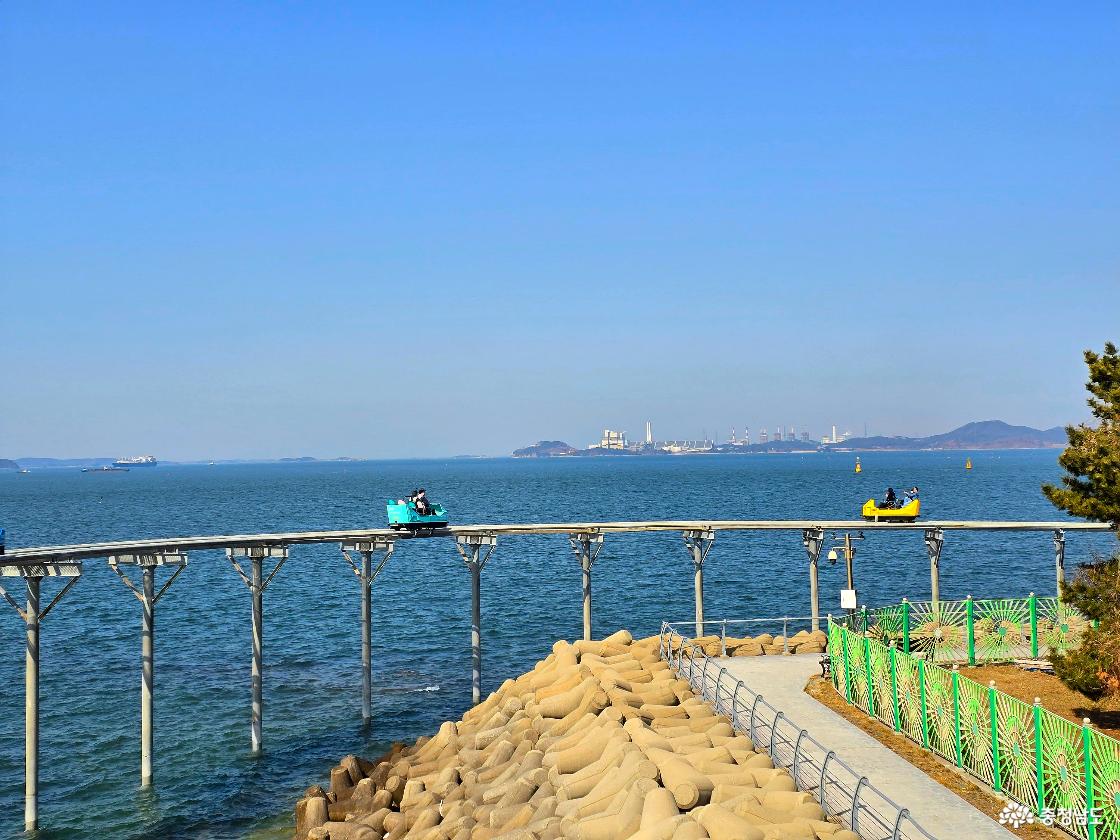 스카이바이크가 바다 위 곡선 레일을 따라 달리는 모습, 아래에는 테트라포드 방파제가 보이고 멀리 산업단지와 섬이 펼쳐진 해안 풍경