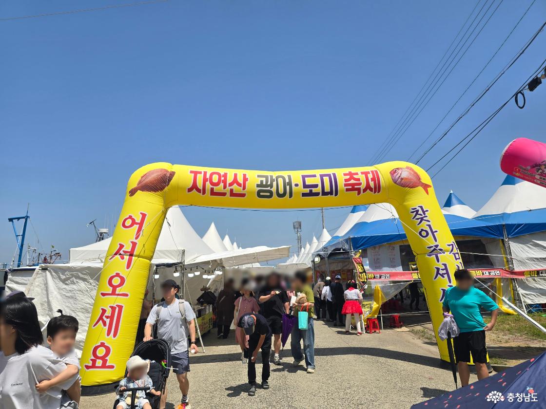 서천 자연산 광어·도미축제 행사장 입구에 노란 대형 아치가 설치되어 있고, 가족 단위 방문객들이 축제장 안으로 들어가고 있는 모습.