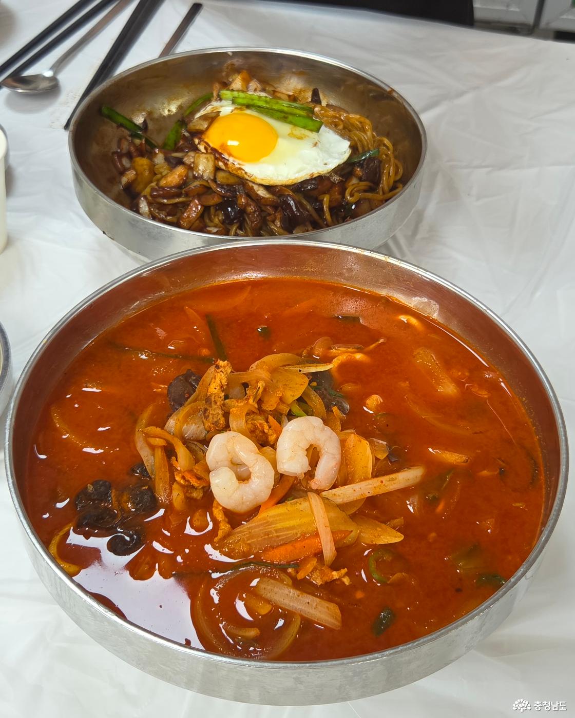 큰 스테인리스 대접에 가득 담긴 붉은 짬뽕과 그 뒤로 계란후라이가 올라간 간짜장이 함께 놓여 있습니다. 짬뽕 국물 위에는 뽀얀 칵테일 새우 두 마리가 나란히 고명으로 얹어져 식욕을 자극합니다.