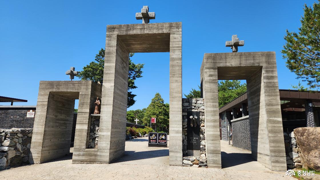 구름 한 점 없이 새파랗고 맑은 하늘을 배경으로 서 있는 충남 당진 솔뫼성지의 주 출입구(정문) 전경. 회색 노출 콘크리트로 정교하게 축조된 세 개의 사각형 문 형태 구조물이 높낮이를 달리하며 나란히 이어져 있다. 가운데 문이 가장 높고 웅장하며, 좌우의 문은 상대적으로 낮게 대칭을 이루는 독특한 비대칭적 조형미를 보여준다. 각 콘크리트 문 구조물의 꼭대기 중심에는 거칠게 다듬어진 돌십자가가 하나씩 우뚝 솟아 있다. 구조물 사이사이는 자연석을 쌓아 올린 석축 기둥으로 연결되어 있으며, 왼쪽 석축 위에는 작은 성인 상이 놓여 있고, 오른쪽 석축 앞에는 두 개의 종이 달린 기념비와 글귀가 새겨진 검은색 석판이 설치되어 있다. 정문 너머 안쪽으로는 푸른 소나무들과 성지 내부의 산책로, 그리고 멀리 작은 하얀색 십자가상이 눈에 들어온니다. 진입로는 깔끔하게 포장된 돌길이며, 정문 왼쪽과 오른쪽 가장자리에는 자연석 돌담과 큰 바위가 배치되어 있다. 가운데 큰 문 아래 안쪽에는 반려동물 출입 금지와 자전거 통행 금지를 알리는 검은색 표지판 두 개가 세워져 있다.