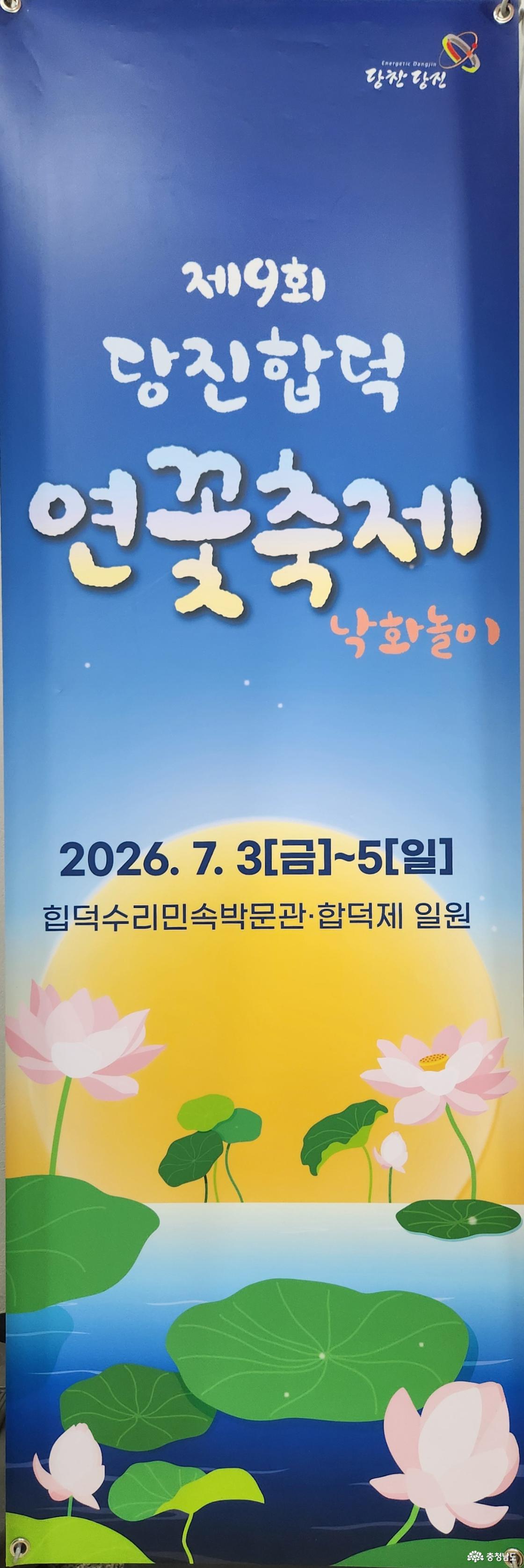 제19회 당진합덕 연꽃축제'를 홍보하는 세로형 배너 포스터 이미지입니다. 상단 텍스트 및 로고: 푸른 밤하늘을 연상시키는 파란색 바탕 상단에 당진시 브랜드 로고인 '당찬당진'이 배치되어 있습니다. 그 아래로 흰색의 감성적인 캘리그래피 글씨체로 '제19회 당진합덕 연꽃축제'라는 메인 타이틀이 크게 적혀 있고, 우측 하단에는 주황색 글씨로 '낙화놀이'가 함께 표기되어 있습니다. 중앙 일정 및 장소: 배너 중간에는 축제 기간인 '2026. 7. 3[금]~5[일]'과 장소인 '합덕수리민속박물관&middot;합덕제 일원'이라는 정보가 짙은 네이비색 글씨로 선명하게 안내되어 있습니다. 하단 일러스트: 배경으로 커다랗고 둥근 보름달 혹은 석양이 노랗고 따스하게 빛나고 있습니다. 그 앞으로 잔잔한 물가에 큼직한 초록색 연잎들과 화사하게 피어난 분홍색 연꽃들이 조화롭게 어우러진 서정적인 일러스트가 그려져 있습니다.