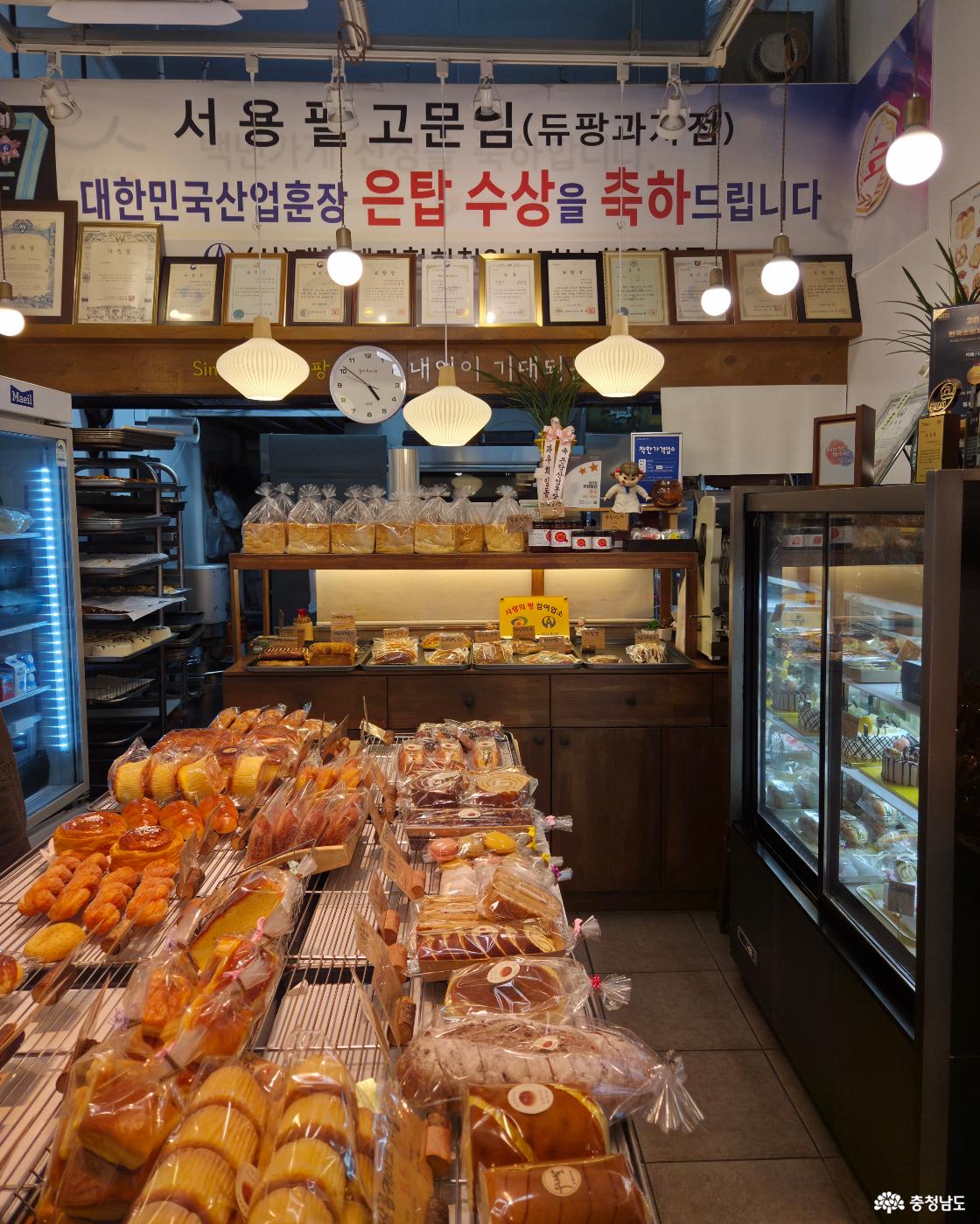 다양한 종류의 빵과 과자들이 진열대에 풍성하게 놓여 있는 빵집 내부 모습입니다. 벽에는 수많은 상장과 '대한민국 산업훈장 은탑 수상'을 축하하는 현수막이 걸려 있습니다.