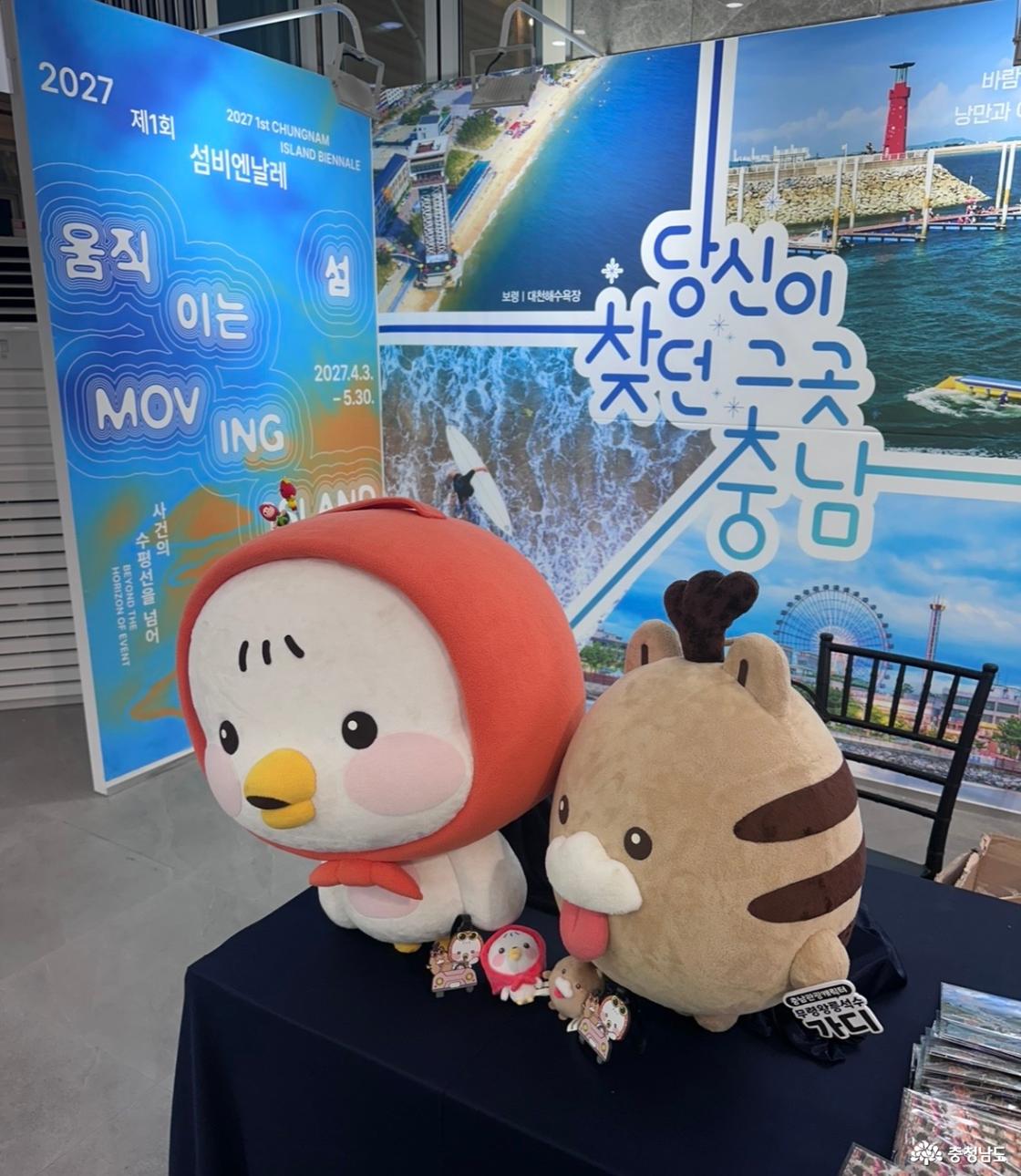 충남 관광 홍보 부스에 놓인 대형 캐릭터 인형과 작은 캐릭터 상품들. 뒤편에 당신이 찾던 그곳 충남 관광 사진과 2027 제1회 섬비엔날레 움직이는 섬 홍보판, 개최 기간 2027년 4월 3일~5월 30일 표시