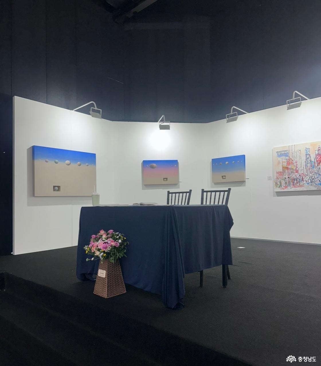 국제아트페어 실내 전시 부스. 흰 가벽에 푸른 하늘과 모래빛 공간, 떠 있는 둥근 형상을 그린 회화 세 점과 다채로운 색의 도시 풍경화가 걸려 있고, 앞에는 남색 천을 덮은 상담 테이블과 꽃바구니
