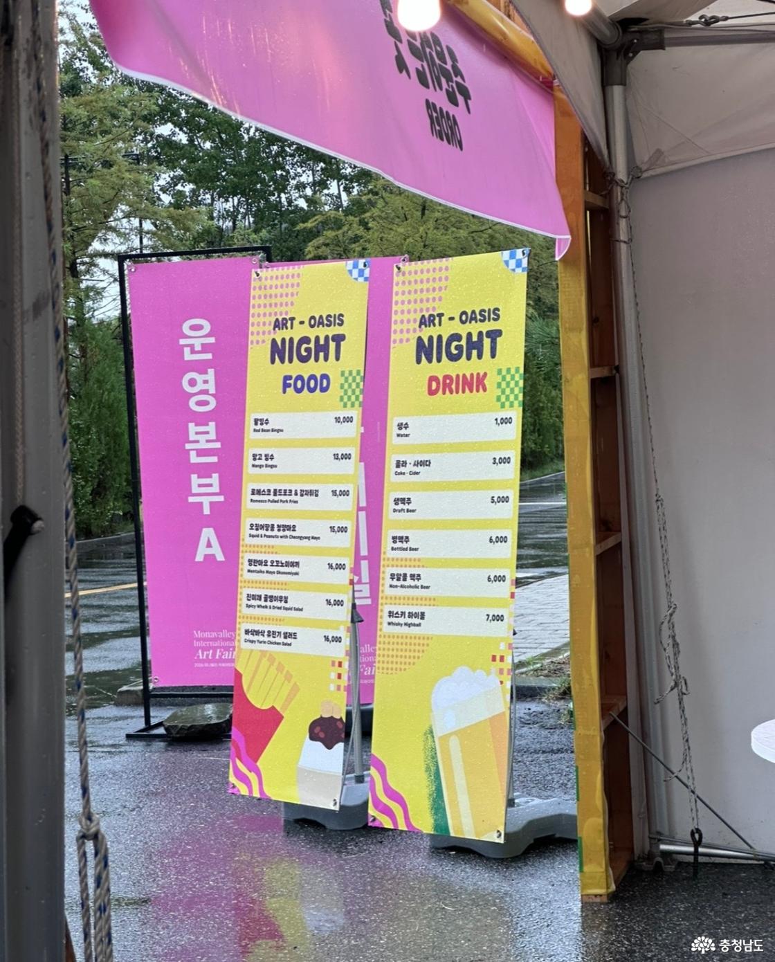 ART-OASIS NIGHT 푸드&middot;음료 메뉴판과 운영본부 A 안내판. 천막 입구에 노랑&middot;분홍색 배너 두 개가 나란히 놓이고, 뒤로 비에 젖은 야외 통로