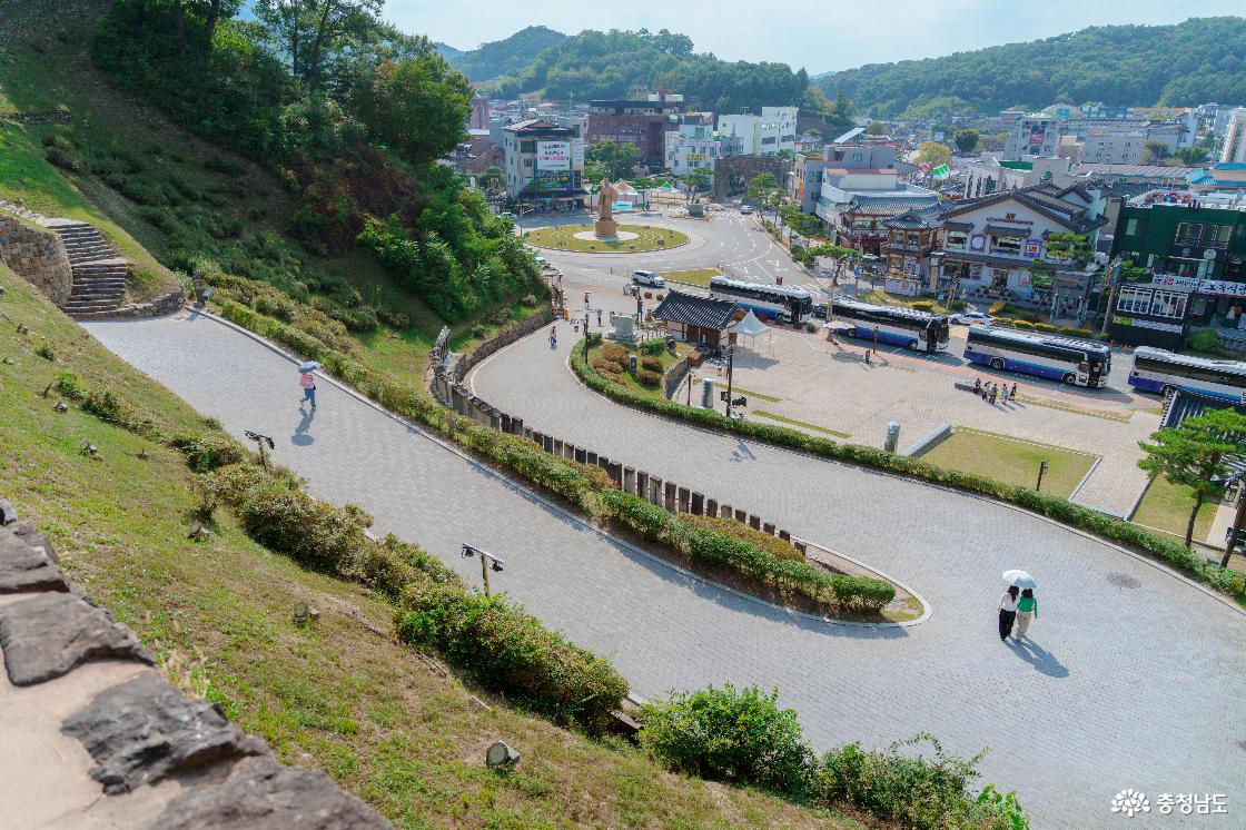 공산성 성벽 위에서 내려다본 굽이진 진입로와 광장. 양산을 쓴 관람객과 줄지어 선 관광버스, 뒤편 공주 시내 건물들.