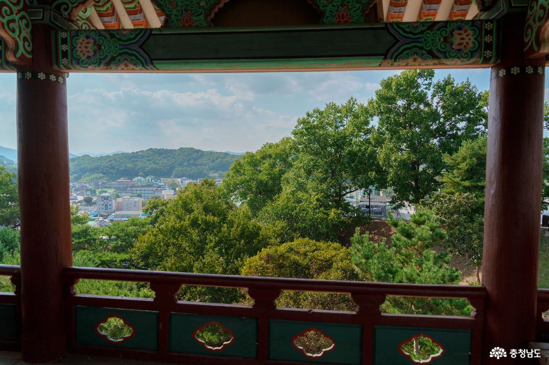 공산정 안에서 기둥과 난간 사이로 바라본 풍경. 가까운 나무들 너머 공주 시내 건물과 낮은 산이 펼쳐진 모습.
