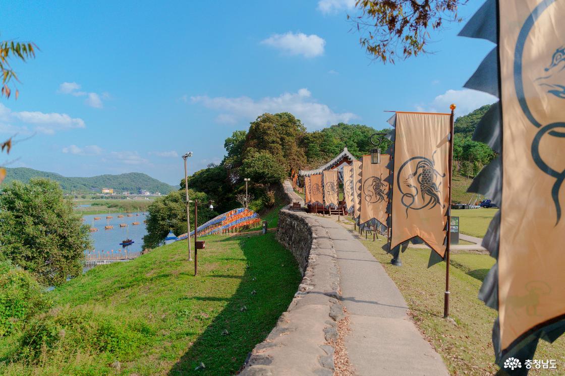 금강을 따라 이어지는 공산성 성곽길과 황색 신수 깃발들. 성 아래 강물 위의 여러 배와 색색의 축제 장식.