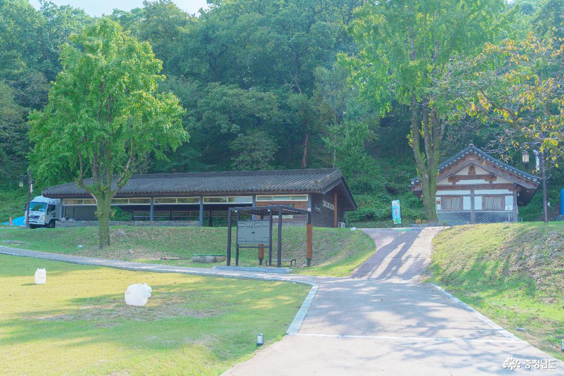 공산성 내부 잔디밭과 산책길 너머의 낮은 기와지붕 건물들. 앞쪽 안내판과 키 큰 나무, 숲이 우거진 비탈.