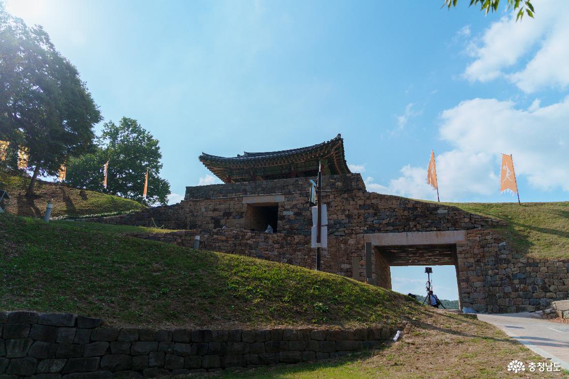 공산성 안쪽 언덕에서 바라본 금서루와 석축 성벽. 문루 아래 사각형 통로와 경사로, 능선 위 황색 깃발들.