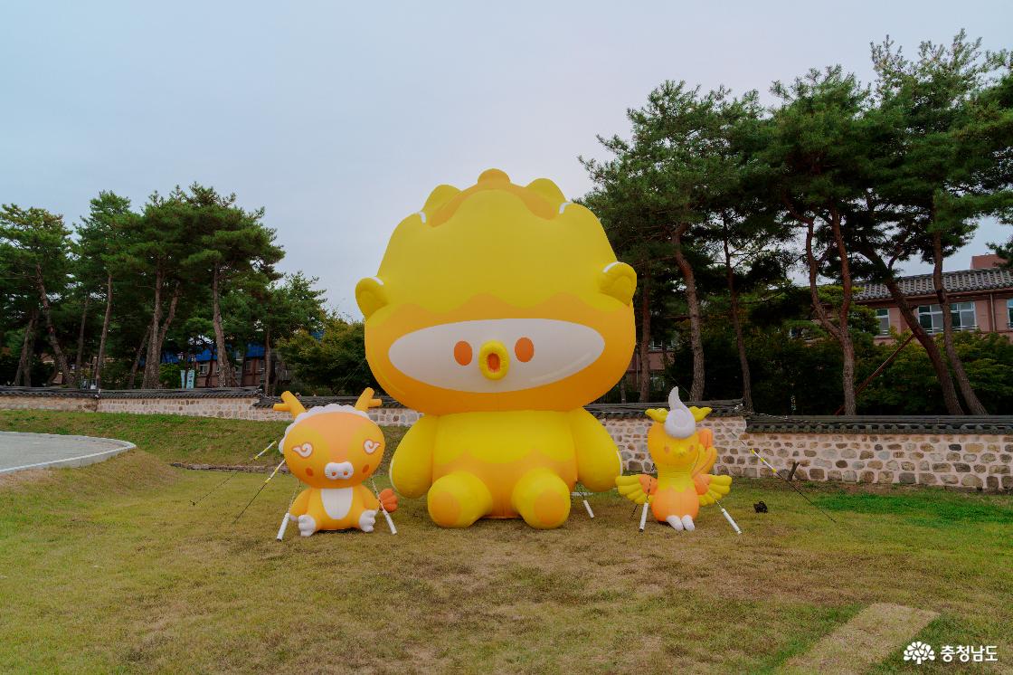 정림사지 잔디밭의 대형 노란 금동이 캐릭터 포토존. 금동이 양옆의 작은 동물 캐릭터 조형물과 뒤편 돌담, 소나무.