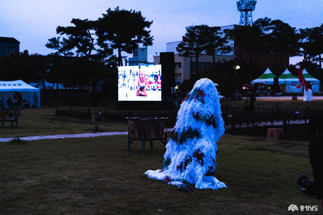 사비천도선포식 잔디밭에서 긴 흰 털 장식으로 온몸을 덮은 탈을 쓰고 공연하는 모습. 뒤편에 큰 화로와 행사 중계 화면.