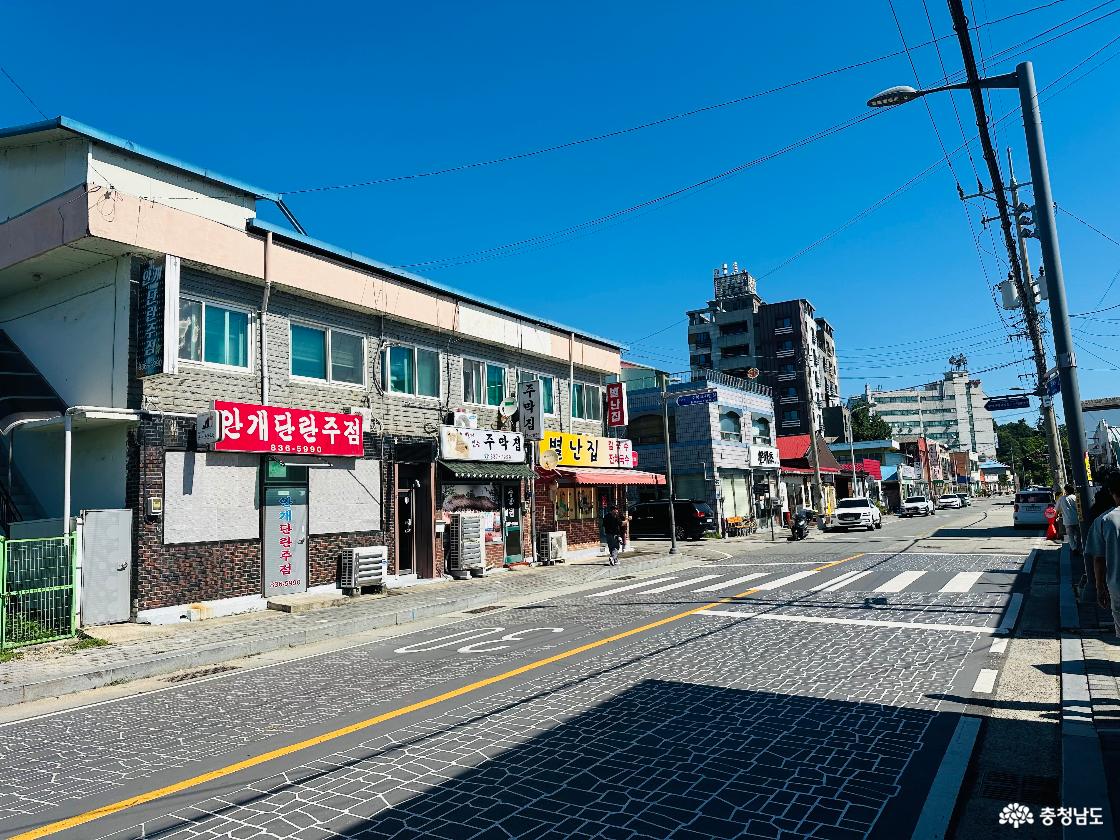 낮은 상가 건물과 오래된 간판이 이어진 부여 규암마을 거리. 도로 위 횡단보도와 길가의 가로등, 전봇대가 보이는 풍경.