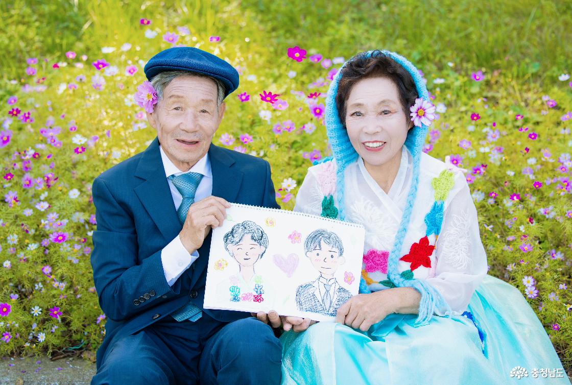 임재호(1943년생)·장복순(1946년생) 부부가 코스모스 꽃밭에서 백경서 학생이 그려준 부부 초상화를 들고 환하게 웃고 있다. 글씨 대신 그림으로 전한 마음이 노부부에게 특별한 추억이 됐다.