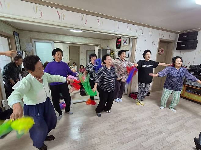 external_image 경로당 실내에서 여러 어르신이 양팔을 벌리고 색색의 천을 흔들며 함께하는 건강체조